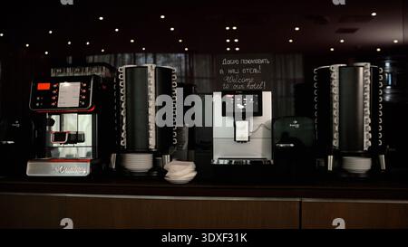 Moderne Selbstbedienungs-Kaffeestation in einer dunklen, eleganten Hotellobby oder einem Konferenzzentrum. Mehrere Espressomaschinen und stapelbare Tassen sorgen für eine einladende Atmosphäre Stockfoto