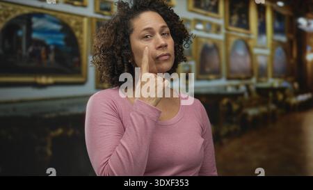 Eine hispanische Frau mittleren Alters zeigt Finger an Wange in einer mit Gemälden gesäumten Gebäudegalerie; Besinnung. Stockfoto