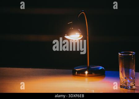 Warme Schreibtischlampe beleuchtet einen Holztisch mit klarem Glas in einem dunklen Innenraum. Minimalistische Nachtszene, ideal für Konzepte wie Fokus, Produktivität usw. Stockfoto