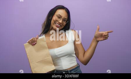 Frau mit brauner Papiertüte zeigt den Finger nach rechts und lächelt mit sichtbarer Hand vor violettem Studio-Hintergrund; verspieltes Selbstvertrauen. Stockfoto