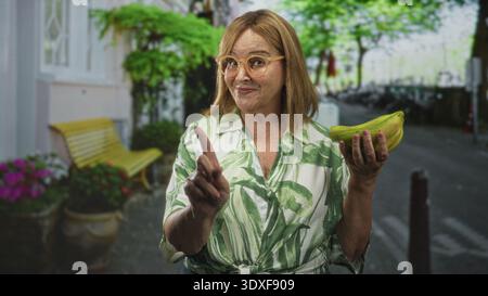 hispanische Frau mittleren Alters mit Brille zeigt mit dem Finger und hält Bananen auf einer Straßenbank mit Topfpflanzen und einer gelben Bank; fröhlich c Stockfoto