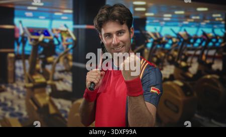 Mann, der Tennisschläger über die Schulter packt und geballte Faust in der Turnhalle mit Laufbändern und Heimtrainer in der Nähe pumpt; Siegesbestimmung. Stockfoto