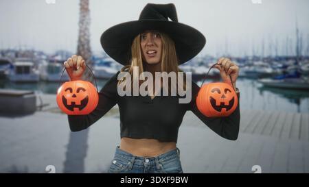 Die junge Frau hält zwei Kürbiseimer an den Griffen, trägt einen Hexenhut mit breiter Krempe und zeigt sichtbare Midriff in einem Studio; verspielter halloween-Spaß. Stockfoto