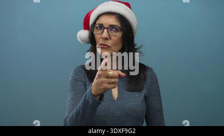 Frau mittleren Alters mit Brille und weihnachtsmann-Hut zeigt mit dem Finger auf die Kamera im Studio mit blauem Hintergrund; Warnung. Stockfoto