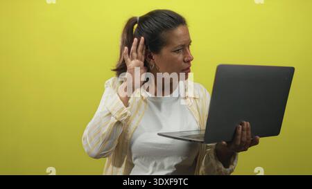 Eine brünette Frau mittleren Alters hält mit einer Hand einen Laptop und schränkt ihr Ohr mit der anderen, während sie im Studio mit gelbem Hintergrund auf den Bildschirm schwindelt Stockfoto