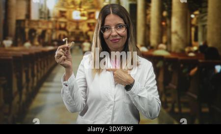 Frau im weißen Mantel, die Hörgerät hält und im Gebäude mit Bänken und Säulen auf sich zeigt; Hörpflegeversicherung. Stockfoto