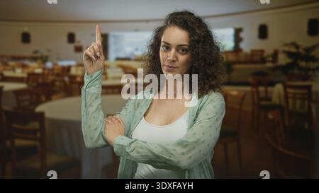 Frau zeigt den Zeigefinger im Speisesaal des Restaurants in der Nähe runder Tische mit umgeklappten Armen und neutralem Blick nach oben; Selbstbewusstsein. Stockfoto