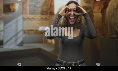 Frau umrahmt Augen mit Händen, zeigt Dekolleté und Brille in einem Gebäude mit Skulpturen, breit lächelnd; Freude verspielte Neugier. Stockfoto