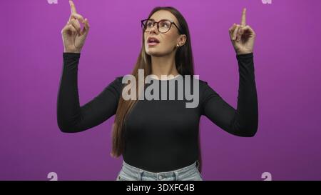 Die junge Frau zeigt beide Zeigefinger nach oben, trägt eine Brille und zeigt Finger und Gesicht in einem Studio; Selbstvertrauen feiern. Stockfoto