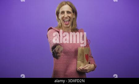 Die lächelnde Frau zeigt mit dem Finger, während sie im Studio eine Papiertüte mit rotem Herzen hält; ein Geschenk, das Freude macht. Stockfoto