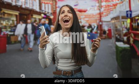 Frau, die Smartphone und Kreditkarte hält, die Hände hochgehoben und den Mund offen hält, auf einem geschäftigen Straßenmarkt; Entlastungszahlung erfolgreich. Stockfoto