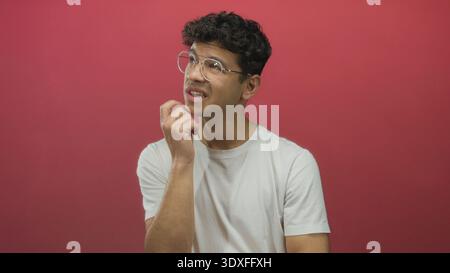 Junger hispanischer Mann mit weißem T-Shirt und runder Brille berührt das Kinn mit nachdenklichem Ausdruck an der roten Studiowand; Zweifel. Stockfoto