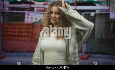 Frau lockiges Haar lächelt mit dem Finger auf die Stirn durch einen orangefarbenen Sicherheitszaun bei Straßenbauarbeiten; Selbstvertrauen. Stockfoto