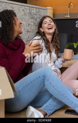 Verschiedene weibliche Freunde sitzen in der Küche und schlürfen von Tassen, die sich an der Arbeitsplatte in der Nähe von Kisten beugen. Gemütlich, Freundschaft, lässig, Zuhause, Lifestyle, fröhlich, warm || Model veröffentlicht Stockfoto