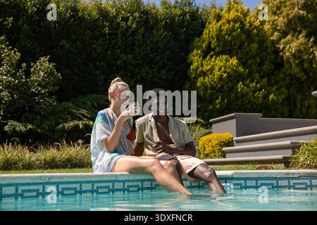 Verschiedene Paare sitzen am gefliesten Poolrand und tauchen die Beine in Wasser und trinken Getränke in Schatten. Luxus, Freizeit, Entspannung, tropisch, Outdoor, lebendig, Lifestyle || Model veröffentlicht Stockfoto
