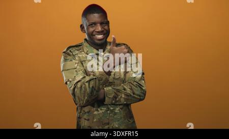 Soldat in Tarnuniform lächelnd mit gekreuzten Armen und erhobenem rechten Daumen im Studio; Stolz. Stockfoto
