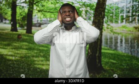 Der Mann bedeckt die Ohren mit Händen im Wald an Fluss, trägt ein weißes Hemd, grimmend und schielend vor Schmerzen; Distress Isolation. Stockfoto