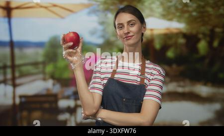 Frau hält Apfel mit Handschuhen auf der Terrasse des Restaurants, trägt Schürze und gestreiftes Hemd; Selbstvertrauen gesunde Ernährung. Stockfoto