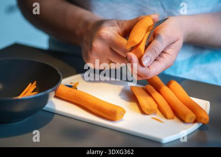 Nahaufnahme der Hände, die frische Karotten mit einem Messer auf einem weißen Schneidebrett schneiden. Mehrere geschälte Karotten liegen neben einer Schüssel in einer Küche. Stockfoto