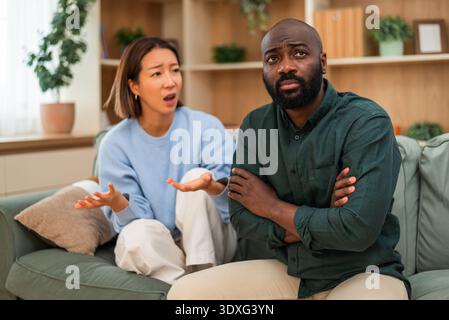Ein multirassisches Paar sitzt zu Hause und streitet sich. Der Mann ist wütend und die Frau redet mit ihm. Stockfoto
