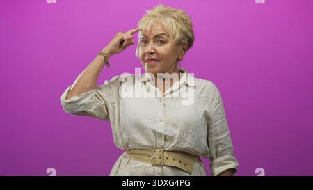 Die ranghöchste hispanische Frau zeigt mit dem Finger auf den Tempel im Studio mit magentafarbenem Hintergrund, trägt ein weißes Leinenhemd und einen beigefarbenen Gürtel; nachdenklich. Stockfoto