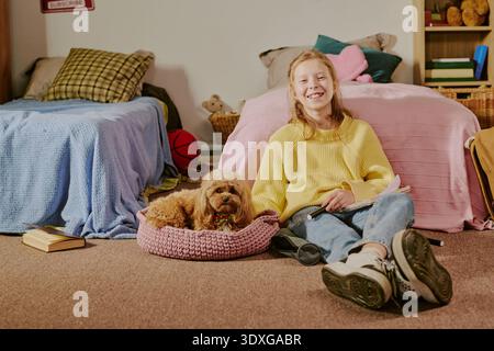 Lächelndes weißes Mädchen sitzt auf dem Boden und liest Buch neben Hund drinnen Stockfoto