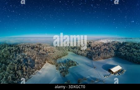 Schnee bedeckt den Boden und die Bäume, während ein Holzbau friedlich in einer weiten, unberührten Winterlandschaft sitzt, die von einem Sternenhimmel beleuchtet wird. Stockfoto