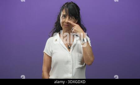 Die Frau zeigt mit dem Finger auf die Nase und lächelt, trägt weiße Bluse und silbernes Armband vor violettem Studiohintergrund; verspielte Wärme. Stockfoto