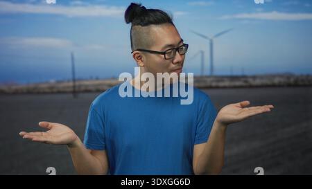 Mann mit den Handflächen auf der Straße, mit blauem T-Shirt und Brille, stehend vor Windmühlenfeld und Himmel; Neugier. Stockfoto