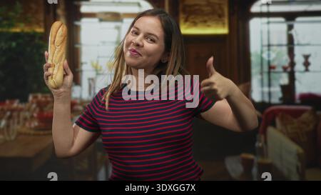 Junge Frau, die ein Baguette mit Handfläche hochhält und Geste im Speisesaal des Hotels in der Nähe von Tisch und Stühlen präsentiert; verspielter Moment. Stockfoto