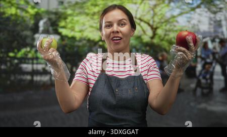 Frau in Denim-Schürze, die einen roten Apfel und einen grünen Apfel mit Handschuhen hält, während sie Obst auf einem Straßenmarkt vergleicht; gesunde Wahl fröhlich. Stockfoto