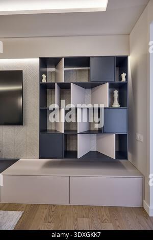 Moderne Wohnzimmerwand mit modularen Regalen, geometrischen Trennwänden, eingebauten Schränken und weicher Hintergrundbeleuchtung über einer niedrigen Tischeinheit. Stockfoto