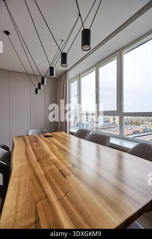 Minimalistischer Essbereich mit langem Holztisch, schwarzen Pendelzylindern an Kabellinien und großen Fenstern mit Blick auf die Skyline der Stadt. Stockfoto