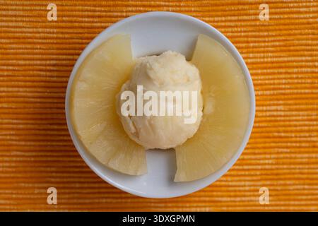 Blick von oben auf einen Löffel Ananassorbet, der in einer weißen Schüssel serviert wird und von frischen Ananasscheiben auf orangefarbenem Hintergrund umgeben ist Stockfoto