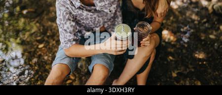 Freunde genießen Drinks im Freien und sitzen in einem sonnigen, entspannten Moment mit einmachgläsern auf dem Boden Stockfoto