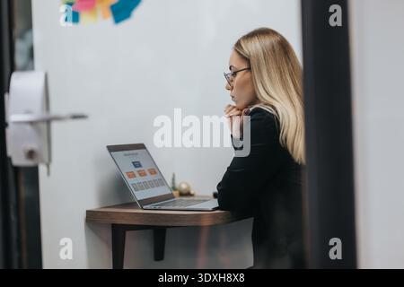 Geschäftsfrau mit Laptop sitzt an einem kleinen Holzschreibtisch in einem modernen Bürobereich Stockfoto