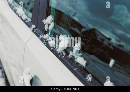 Schneehaufen auf dem Seitenfenster eines weißen Autos, das während eines Wintersturms in einer kalten, dunklen Jahreszeit in der Nähe von Häusern geparkt wurde. Die Szene zeigt die Auswirkungen von starkem Schnee Stockfoto