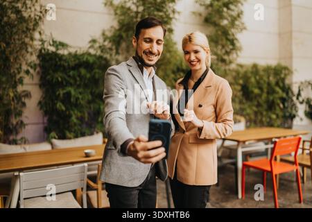 Zwei Geschäftspartner posieren für ein Selfie in einem Café im Freien, lächelnd und elegant gekleidet. Stockfoto