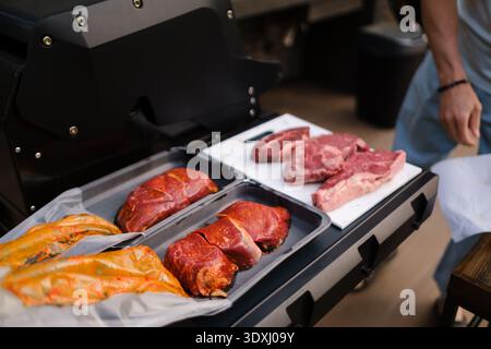 Jemand bereitet verschiedene Fleischsorten auf einem Grill zu. Verschiedene Rindfleisch- und Lachsstücke sind ordentlich angeordnet. Es ist ein sonniger Nachmittag, und die Umgebung ist Stockfoto