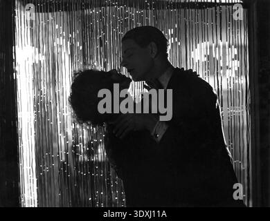 Dieses emotional aufgeladene still aus dem Stummmelodram „Downhill“ aus dem Jahr 1927 fängt die Schauspieler Robin Irvine (rechts) und Annette Benson (links) in leidenschaftlicher Umarmung ein. Unter der Regie von Alfred Hitchcock zeigt das Schwarzweißbild einen dramatischen Moment mit starker Hell-Dunkel-Beleuchtung, die die Konturen ihrer Gesichter und Hände vor einem schimmernden, vertikalen Hintergrund betont. Stockfoto