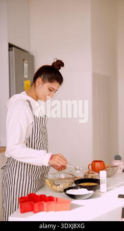 Junge Frau in Schürze, die Eier in eine Glasschale knackt, während sie beiläufig kocht Stockfoto