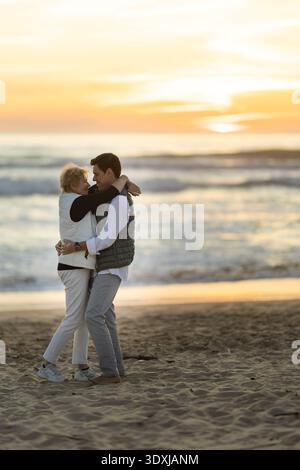 Ältere Frau und Mann, die sich bei Sonnenuntergang am Strand umarmen Stockfoto