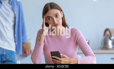 Frau sitzt am Tisch mit Smartphone und zeigt Zeichen von Stress in der Küche Stockfoto