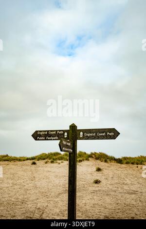 Ein hölzerner Wegweiser auf einem sandigen Pfad weist in entgegengesetzte Richtungen entlang des „England Coast Path“ und „Public Footpath“ mit Dünengras und bewölktem Himmel Stockfoto
