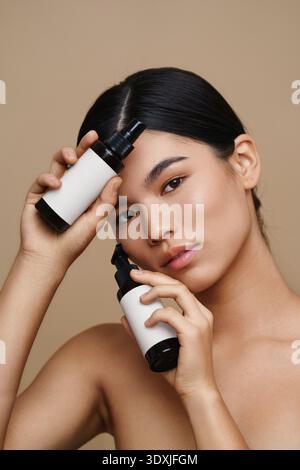 Eine ruhige Frau hält zwei Kosmetikflaschen mit leeren Etiketten, perfekt für Mockups von Kosmetikprodukten. Der gleichmäßige Hautton und das minimalistische Design strahlen aus Stockfoto