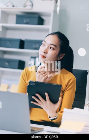 Asiatische Frau arbeitet am Laptop am Schreibtisch. Sie lächelt glücklich, während sie auf der Tastatur neben dem Fenster mit Jalousien schreibt. Professionelle Frau arbeitet im Geschäft Stockfoto