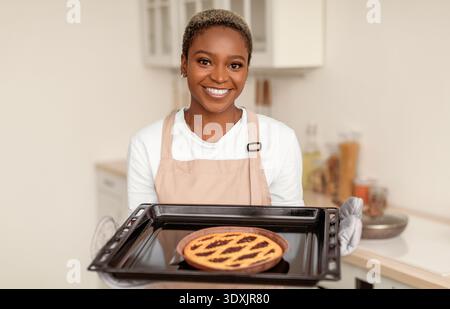 Eine junge Frau hält einen frisch gebackenen Kuchen in einer Küche, während sie die Kamera freundlich anlächelt Stockfoto