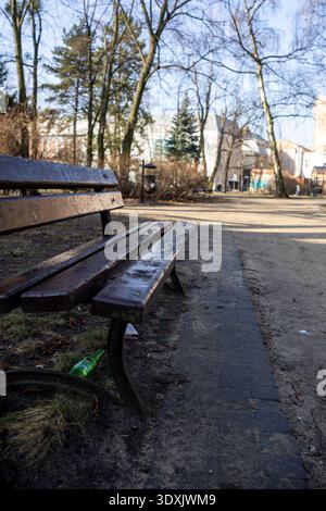 Ein stimmungsvoller und melancholischer Schuss einer nassen, leeren Holzbank in einem vernachlässigten Stadtpark. Der Boden um die Bank herum ist voller Müll, einschließlich eines Di Stockfoto