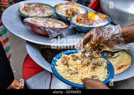Blick auf Dampfplatten und Schüsseln, die hoch mit Reis und Fleisch gefüllt sind, ein Verkäufer, der Speisen im belebten Chawkbazar, Dhaka, Dhaka Division, Bangladesch serviert. Stockfoto