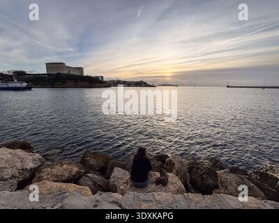 Vieux Port (Alter Hafen) Marseille, Frankreich. Die Person sitzt auf Felsen und beobachtet den Winteruntergang über dem Mittelmeer in der Nähe des Hafeneingangs. Stockfoto